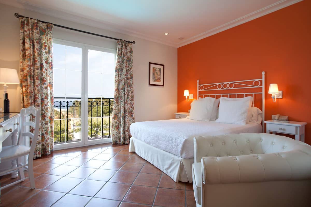 A hotel room with a bed, two bedside tables, a chair, a balcony with a railing, and a view of greenery.