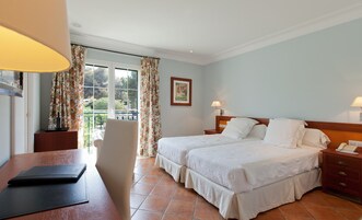 A hotel room with two beds, a desk, a chair, and a view of the outdoors.