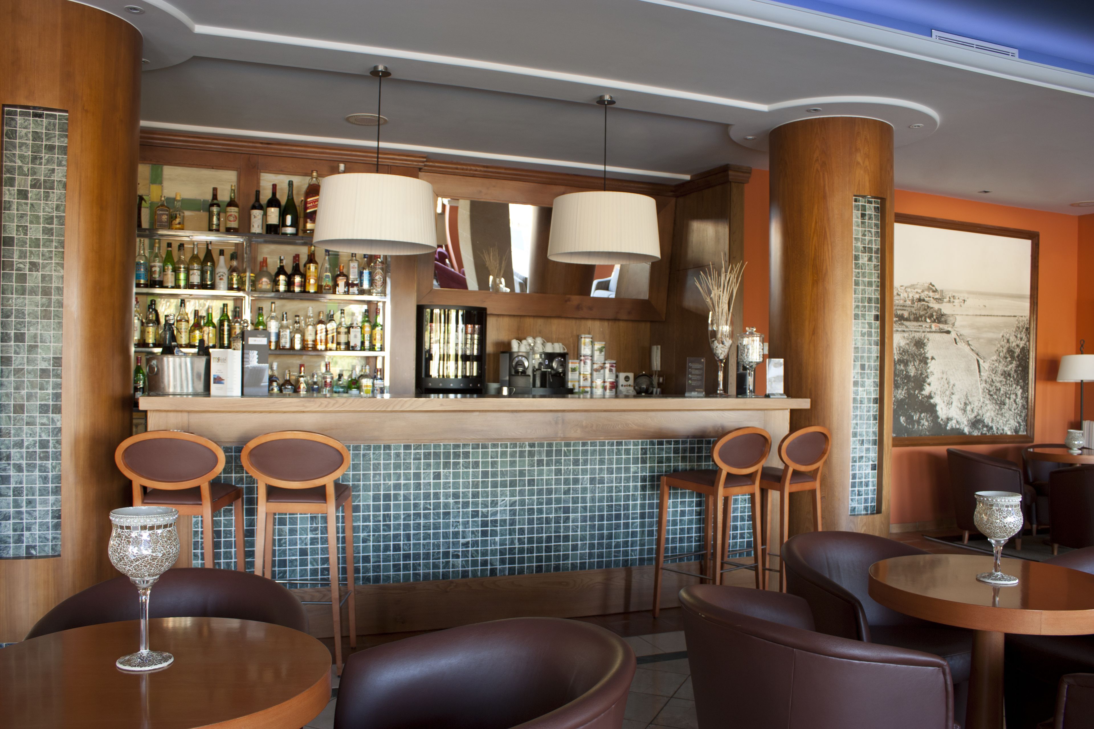 Un espace bar agrémenté d’un mur recouvert de carreaux, d’un comptoir en bois et de différents types de sièges, notamment des tabourets de bar et des tables rondes.