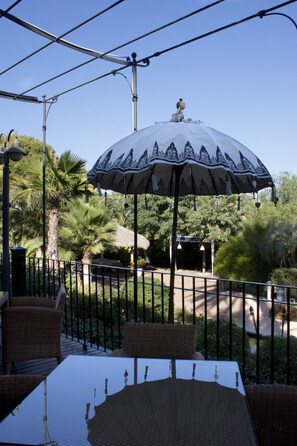 An outdoor dining area with a table and chairs, a decorative umbrella, and a view of a garden with palm trees. - Hotel Les Rotes (Denia)