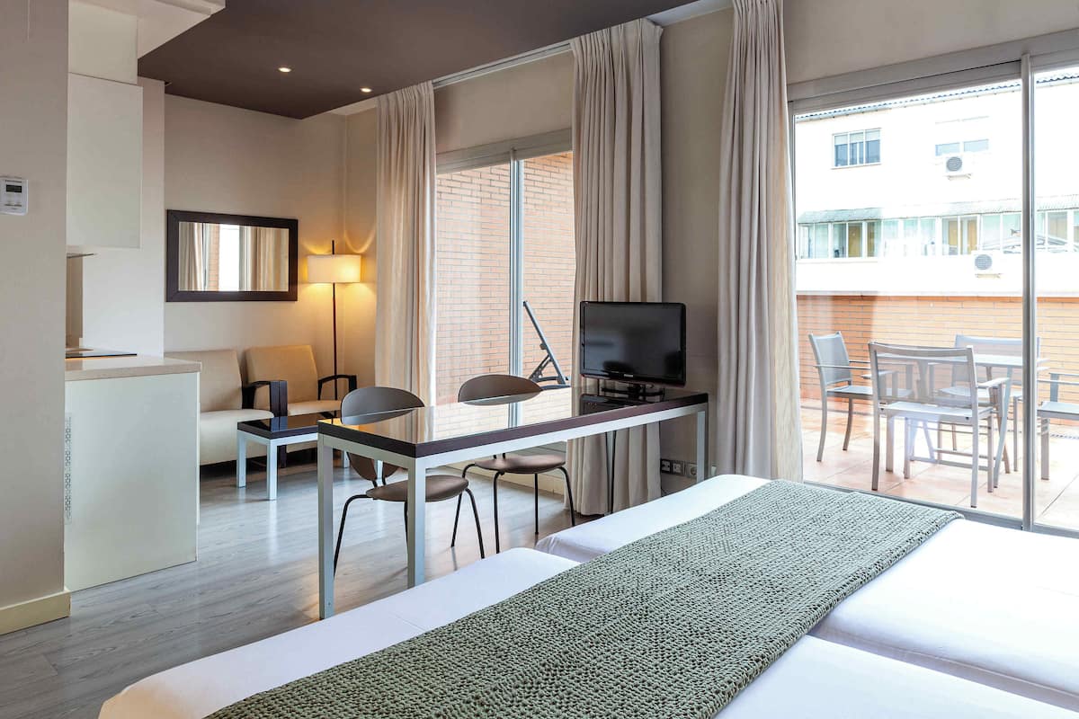 A hotel room with a bed, a desk with a TV, a chair, a sofa, and a balcony with chairs.