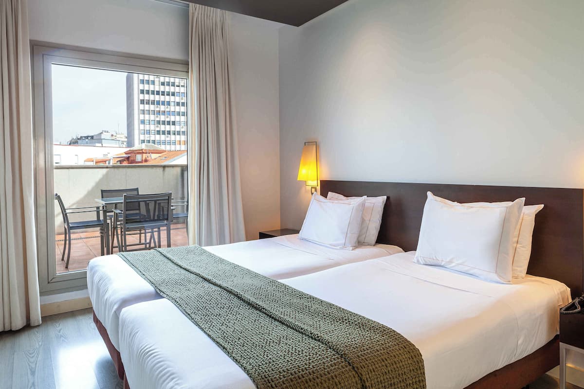 A hotel room with two beds, a balcony with a table and chairs, and a city view.