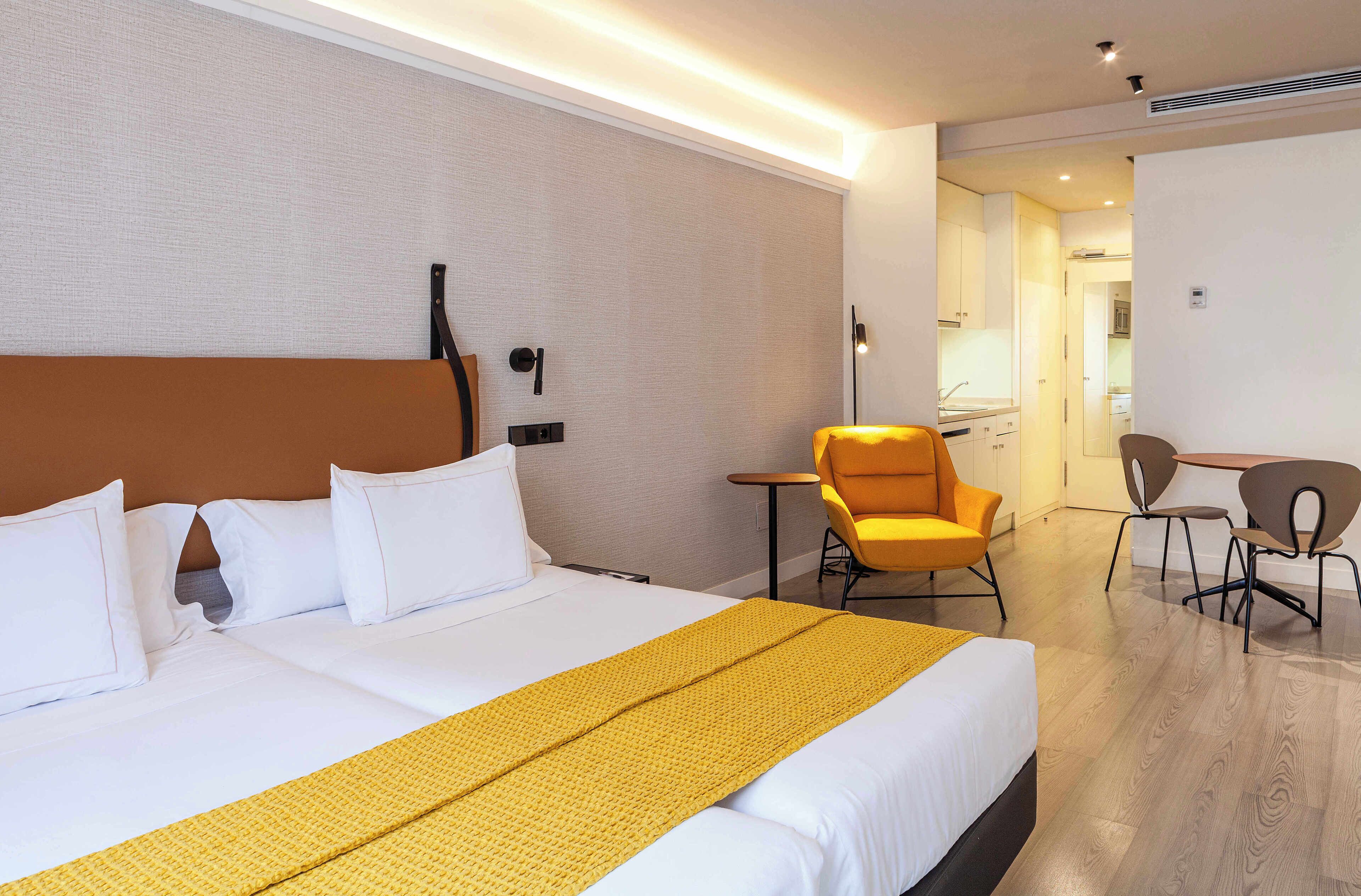 Habitación de hotel con una cama grande, una mesita, dos sillas y una kitchenette al fondo.