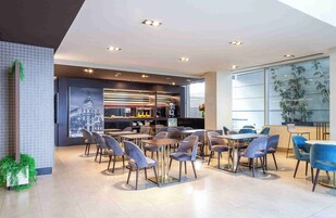 A modern restaurant with a bar area, seating with tables and chairs, and large windows with plants.