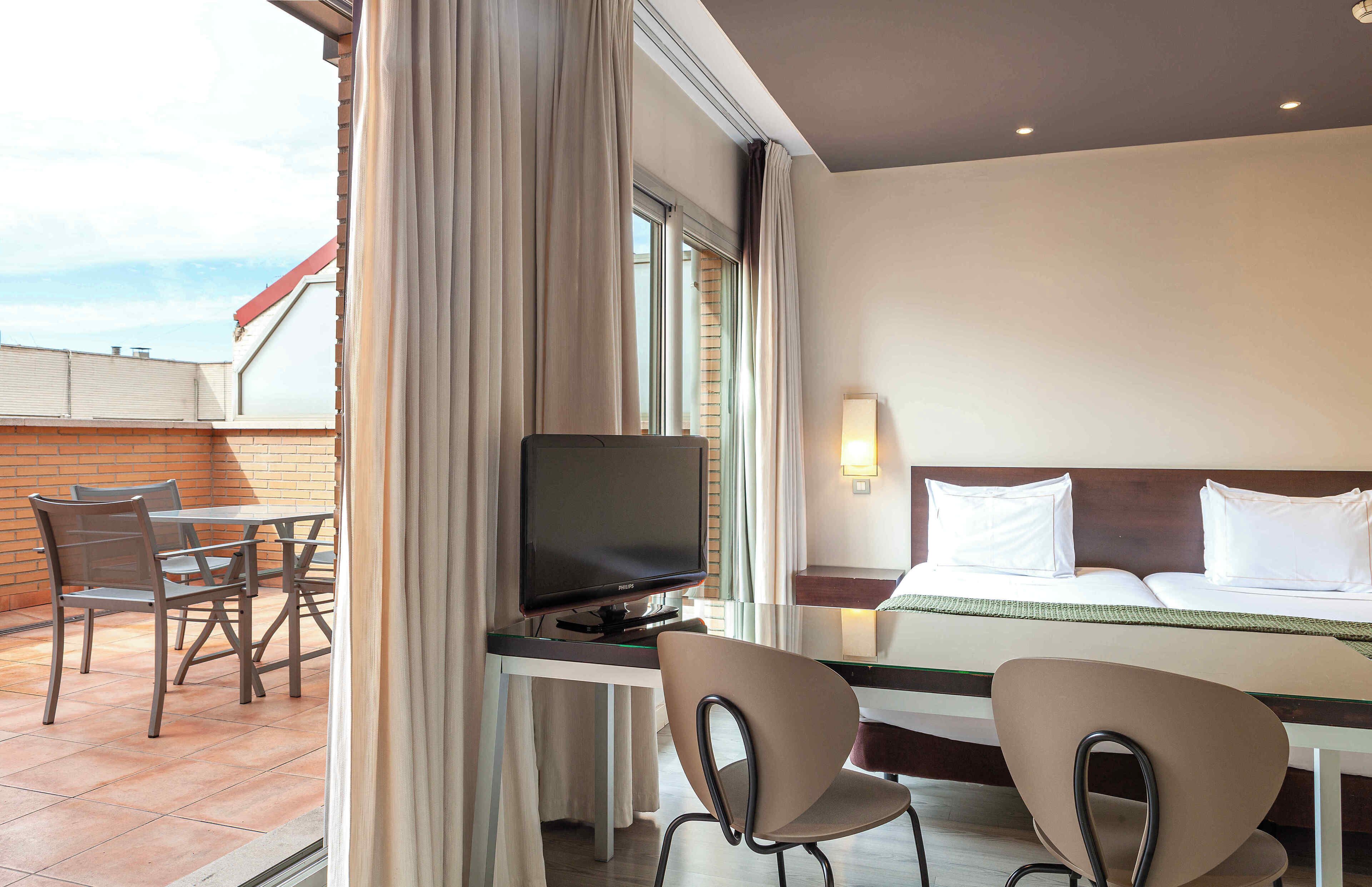 A hotel room with a bed, a desk with a television, two chairs, and a window with curtains.