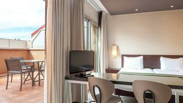 A hotel room with a bed, a desk with a television, two chairs, and a window with curtains.