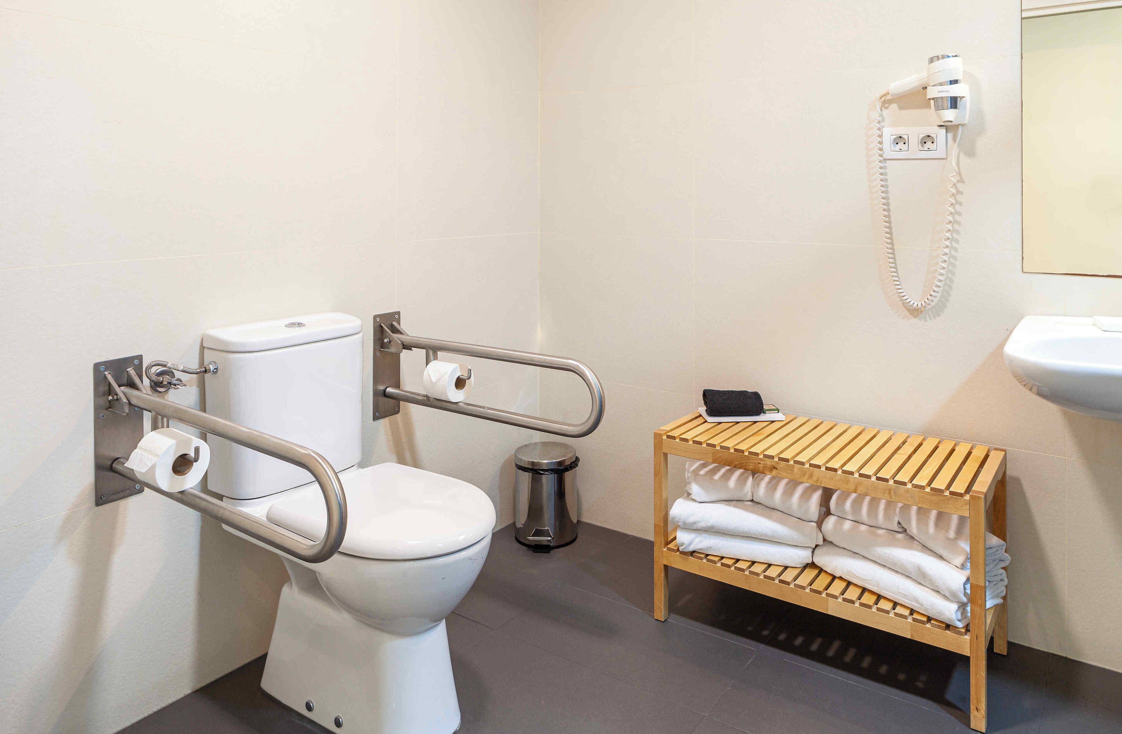 Une salle de bain moderne équipée de toilettes, de barres d’appui, d’une poubelle, d’un porte-serviettes et d’un sèche-cheveux.