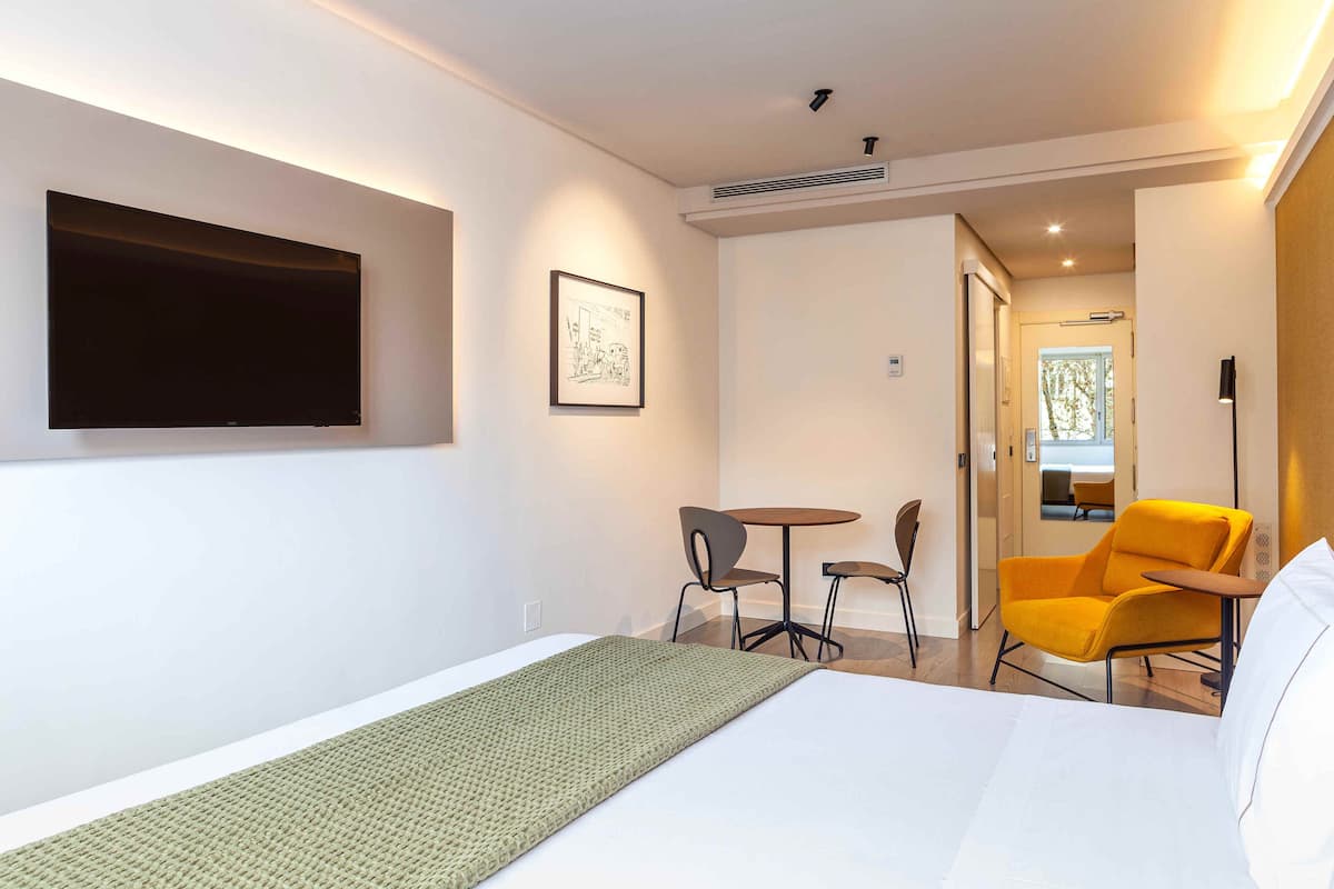 A hotel room with a bed, a flat-screen TV mounted on the wall, a small table with two chairs, and a bright yellow armchair.