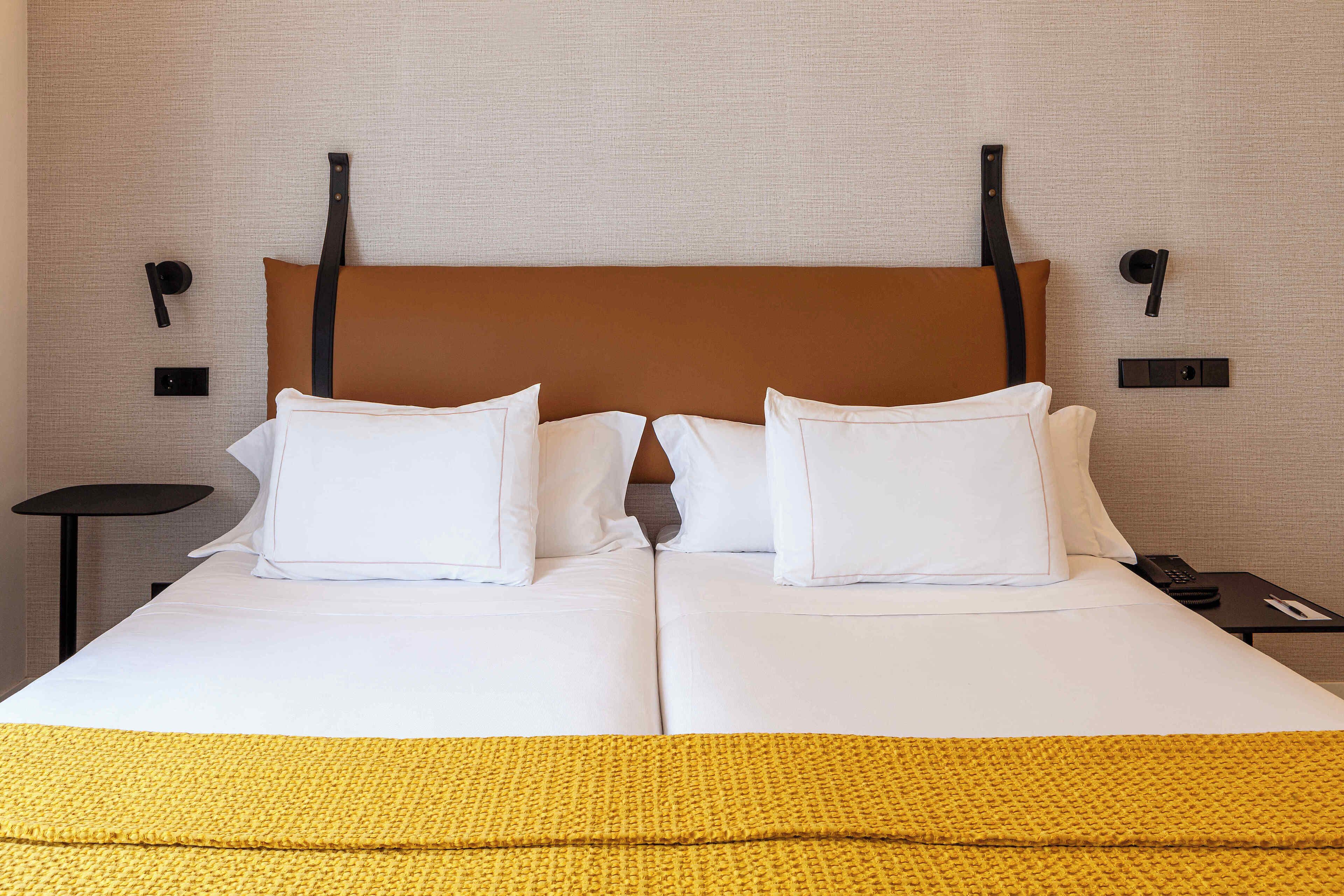Una cama doble con ropa de cama blanca y un cabecero marrón, una mesita de noche pequeña con un control remoto y una lámpara de pared.