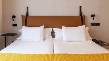 Una cama doble con ropa de cama blanca y un cabecero marrón, una mesita de noche pequeña con un control remoto y una lámpara de pared.