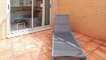 A sunlit patio with a metal lounge chair, a sliding glass door leading to a kitchen area, and a brick wall.