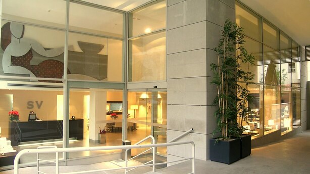 A modern hotel lobby with glass walls, a reception desk, and a large potted plant. - Suites Viena Plaza de España (Madrid)