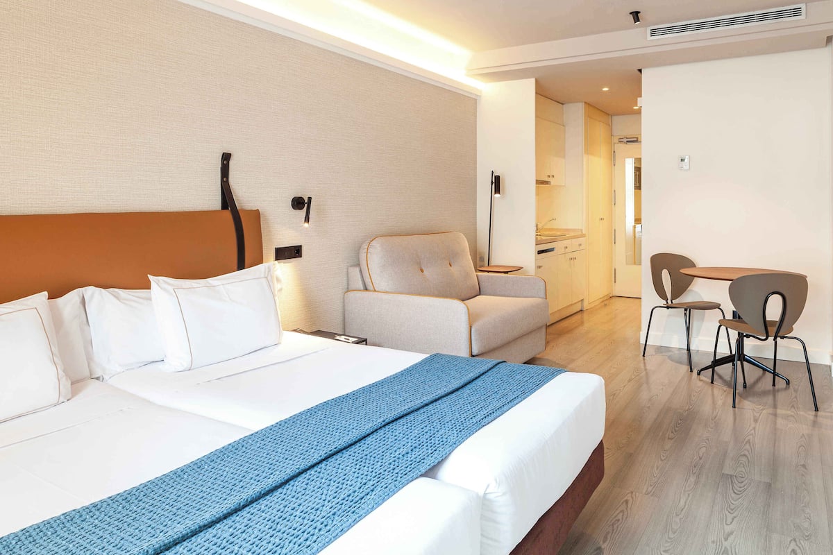 A hotel room with a bed, a chair, a desk, and a kitchenette.