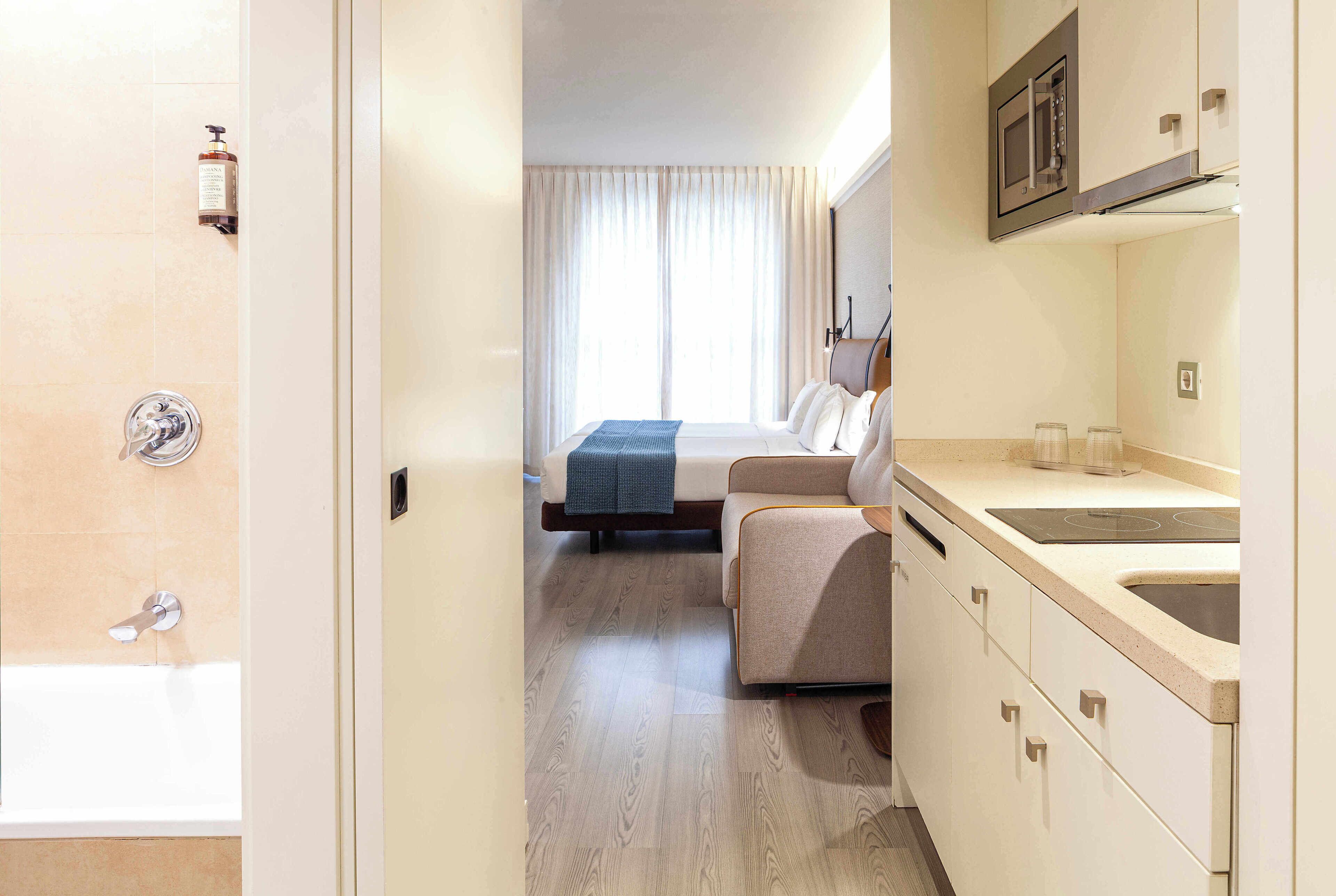 A hotel room with a shower, a bed, a sofa, a kitchen area, and a window with curtains.