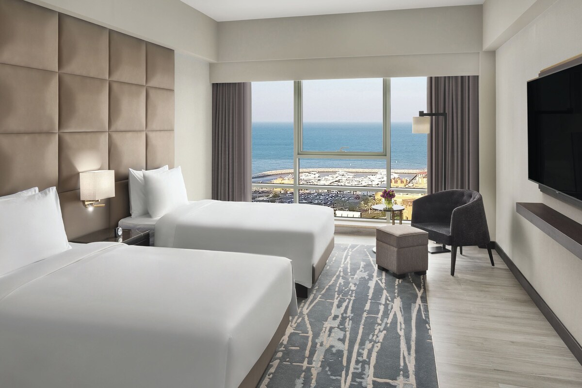 A hotel room with two beds, a TV, a chair, and a view of the beach.