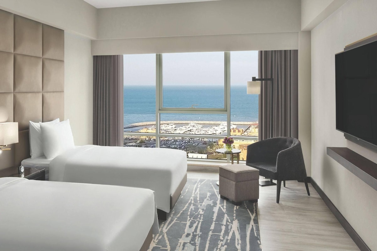 Habitación de hotel con dos camas, televisión, una silla y vista a la playa.