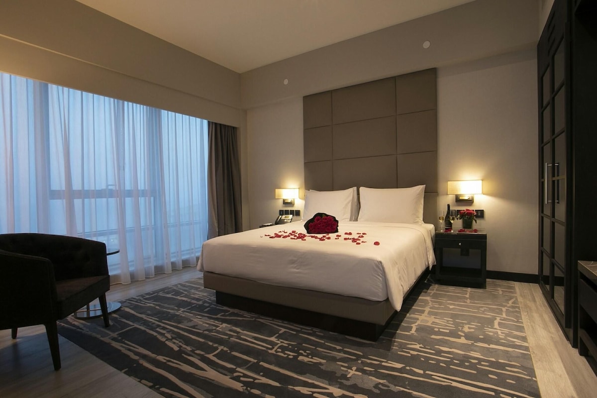 Habitación de hotel con una cama grande, dos mesitas de noche con lámparas, un sillón oscuro y un ventanal con cortinas traslúcidas.