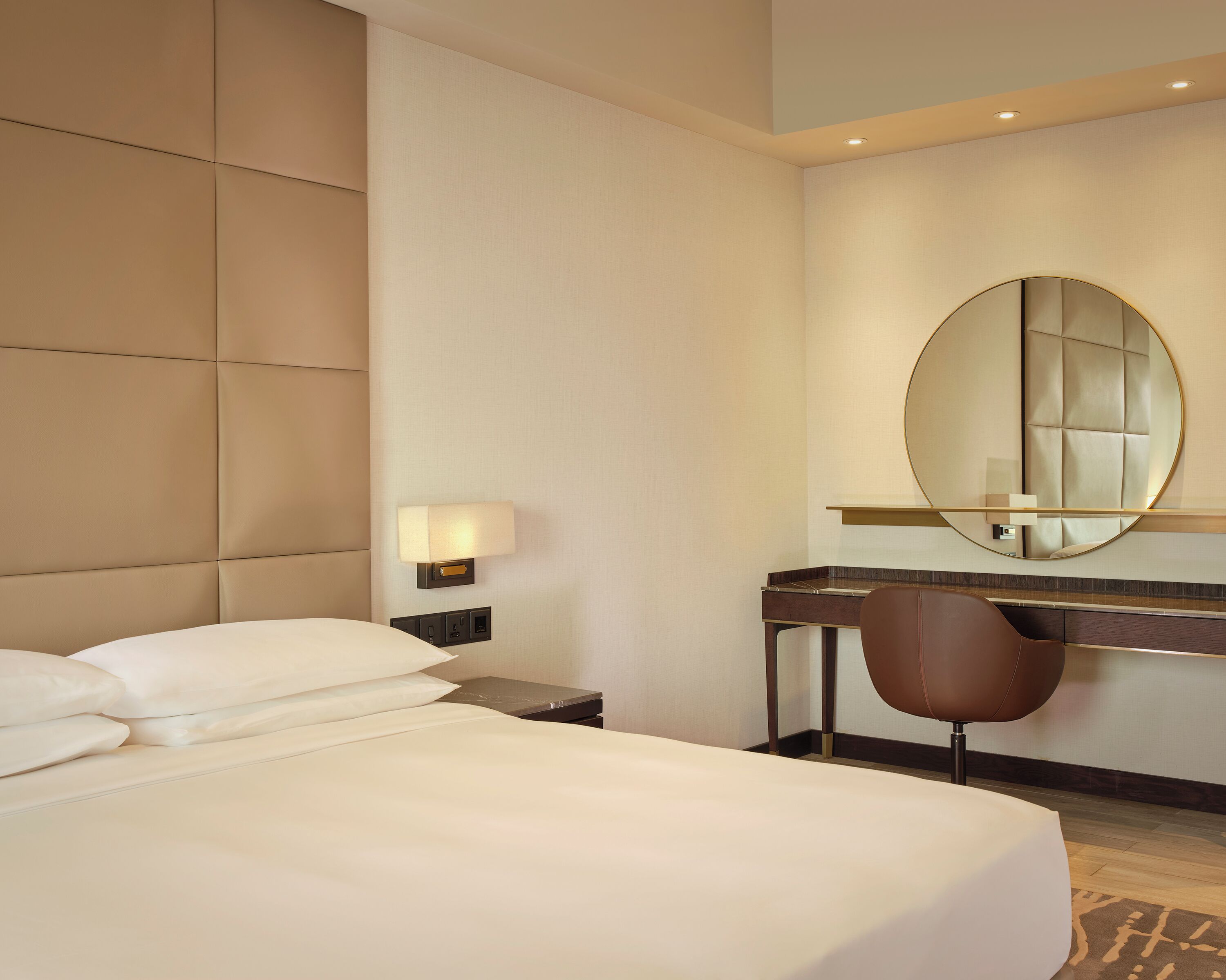 A hotel room with a bed, a desk, a chair, a large mirror, and a wall-mounted lamp.