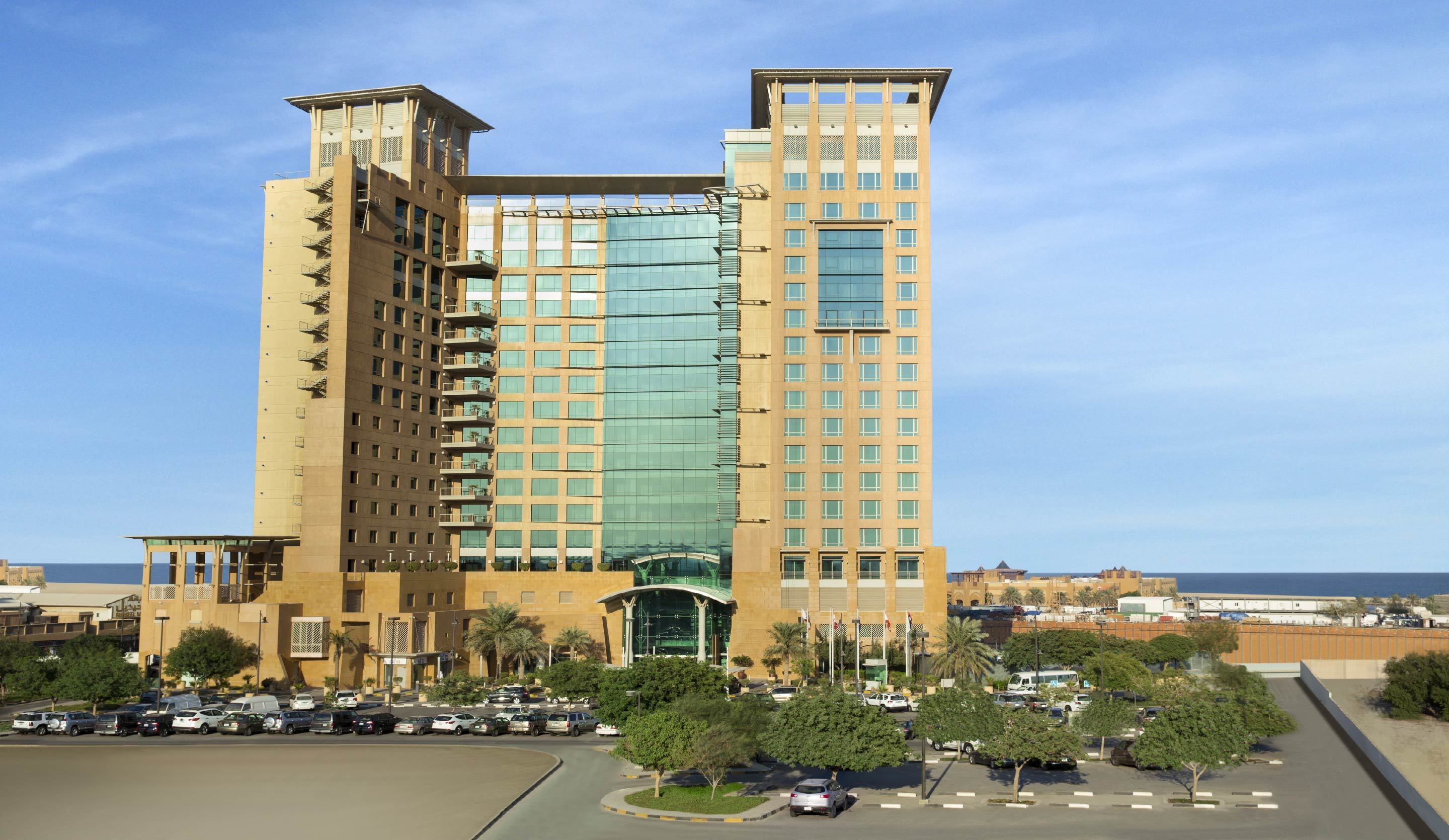 A modern hotel with a central glass facade, multiple balconies, and a parking lot with cars and palm trees.