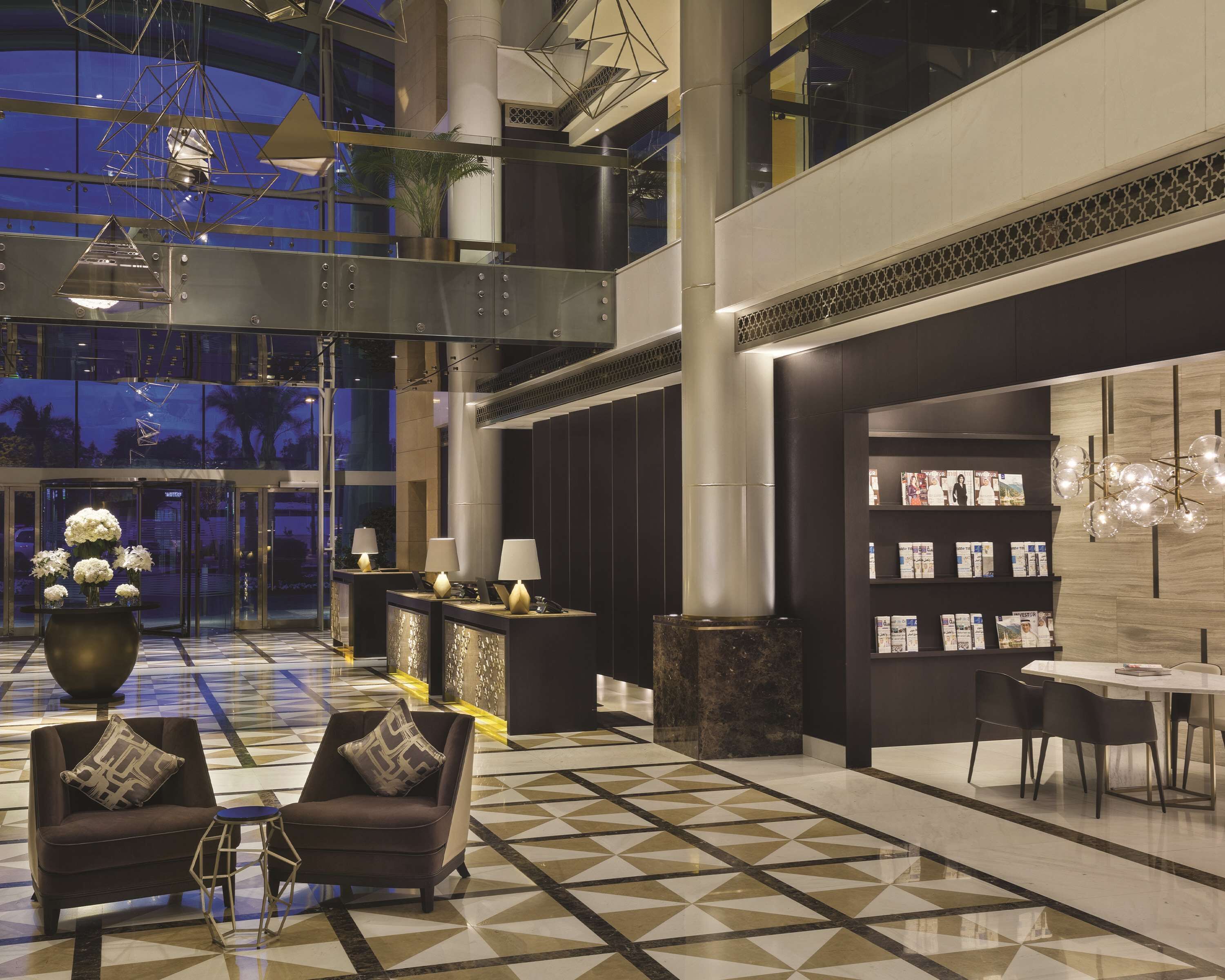 A spacious lobby with a geometric patterned floor, a reception desk with a lamp, a seating area with a sofa and chairs, and a wall with a display of magazines.
