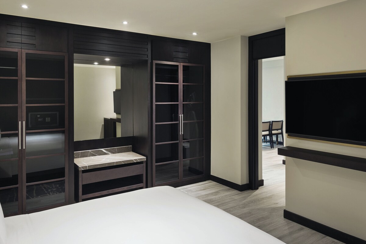 Una habitación de hotel moderna con una cama grande, un armario empotrado con puertas de cristal, un televisor de pantalla plana en la pared y una pequeña zona de estar con una mesa y sillas.