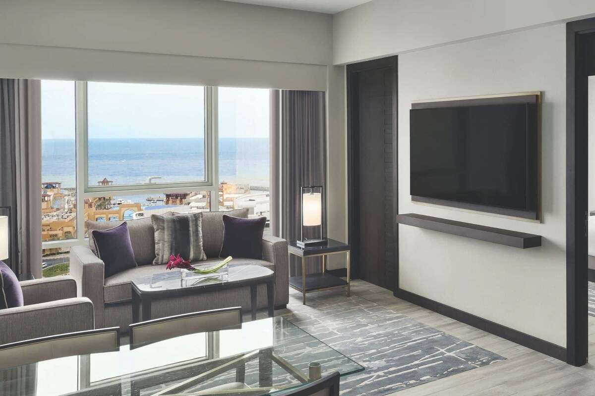 Una habitación de hotel moderna con un ventanal que ofrece vistas a la ciudad, un televisor de pantalla plana montado en la pared, una cama con ropa blanca y una zona de estar con una mesa de vidrio y cojines.