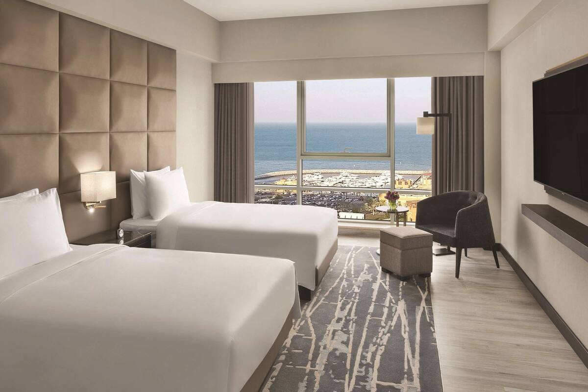 Quarto de hotel com duas camas, televisão, uma cadeira e vista para a praia.