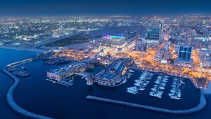 An aerial view of a marina with boats docked, surrounded by buildings and a cityscape at night. - Hyatt Regency Al Kout Mall (Fahaheel)