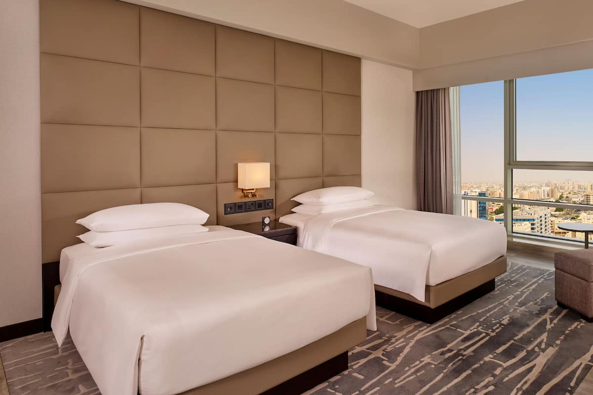 Quarto de hotel com duas camas, uma mesinha de cabeceira, uma luminária e vista para a cidade.