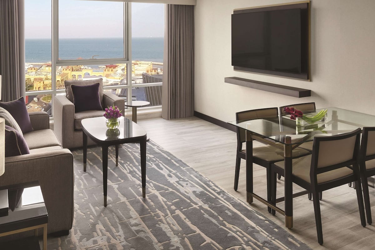 Quarto de hotel moderno com uma janela grande que oferece vista para a cidade, uma televisão de tela plana na parede, uma área de jantar com mesa de vidro e um sofá com um vaso decorativo.
