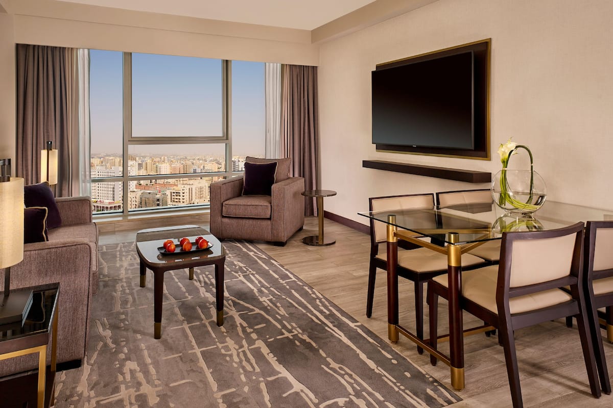 Quarto de hotel moderno com uma grande janela que oferece vista para a cidade, uma TV de tela plana na parede, uma mesa de jantar de vidro com cadeiras, um sofá e uma mesa de centro com frutas.