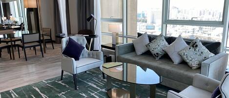 A modern hotel room with a sofa, armchairs, a dining area, and large windows offering a city view.