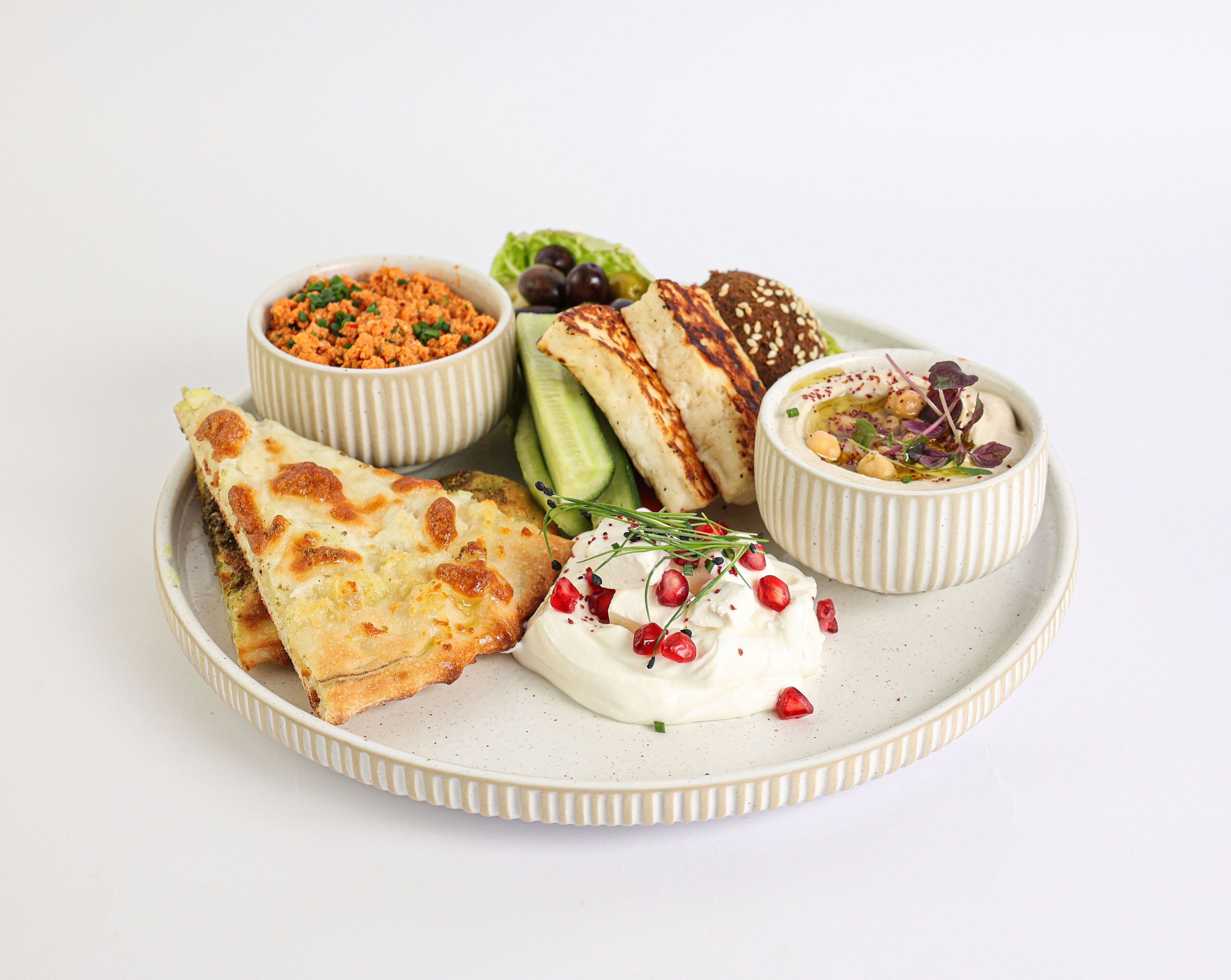 Un plato con diversos alimentos, incluyendo un pastel horneado, un tazón de hummus, una ensalada, un yogur con semillas de granada y un tazón de aceitunas.