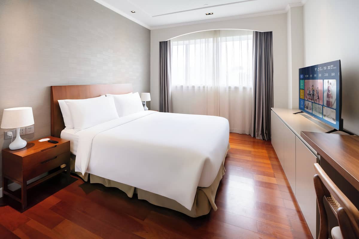 A hotel room with a large bed, a bedside table with a lamp, a TV, and a wooden floor.
