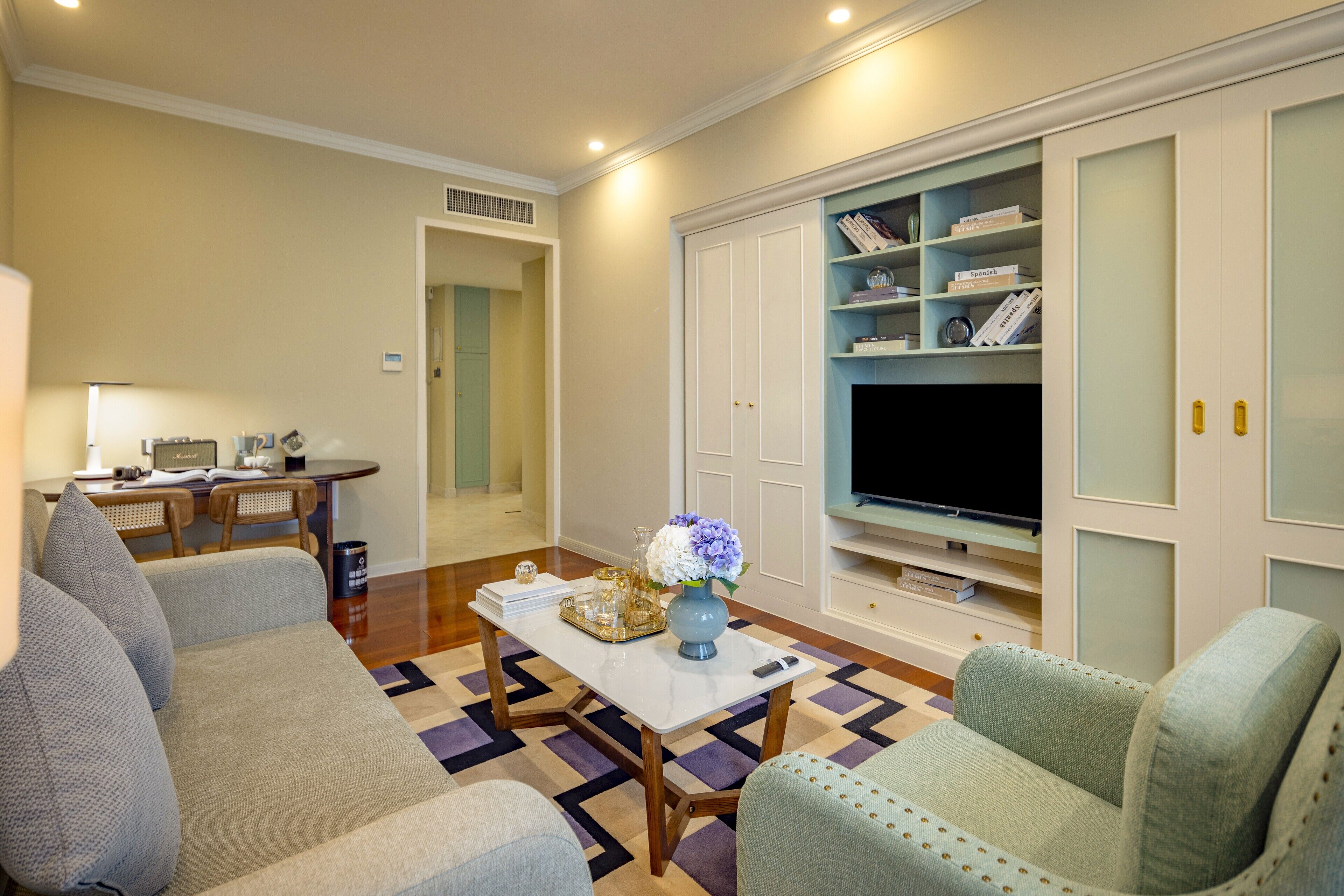 A living room with a sofa, a coffee table, and a TV. There is a bookshelf with books and decorative items.
