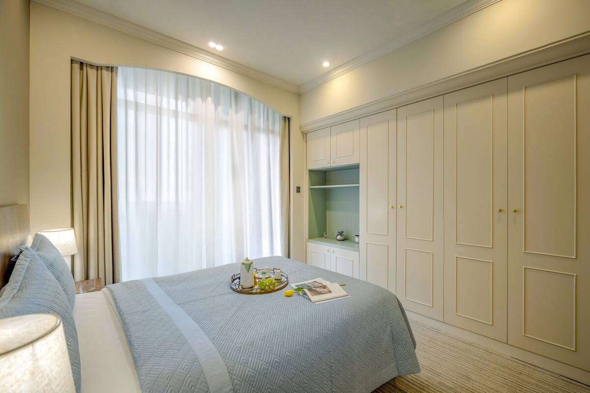 A hotel room with a bed, a nightstand, a lamp, a window with curtains, a wardrobe, and a small shelf with decorative items.