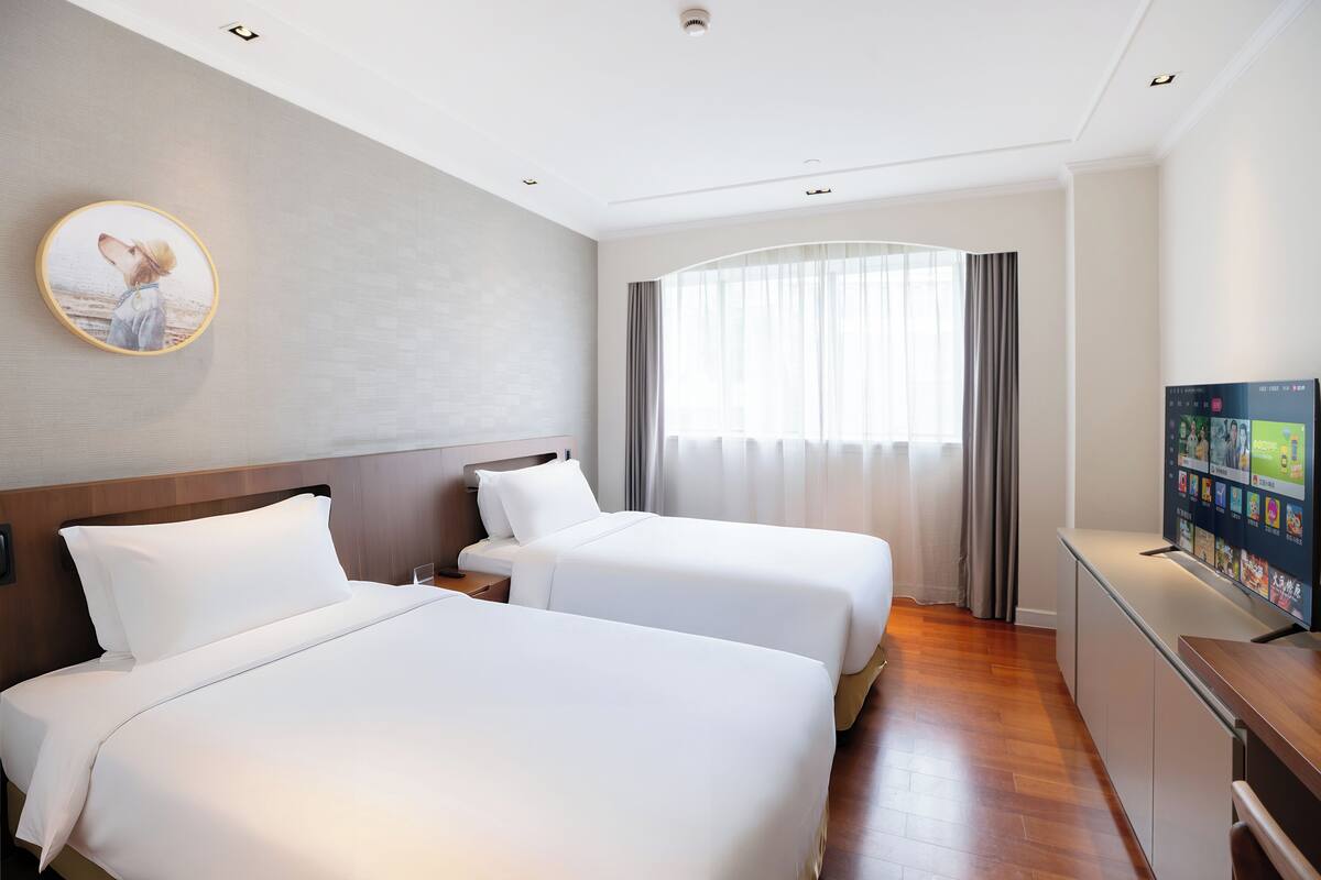 A hotel room with two beds, a TV, a large window with curtains, and a painting on the wall.