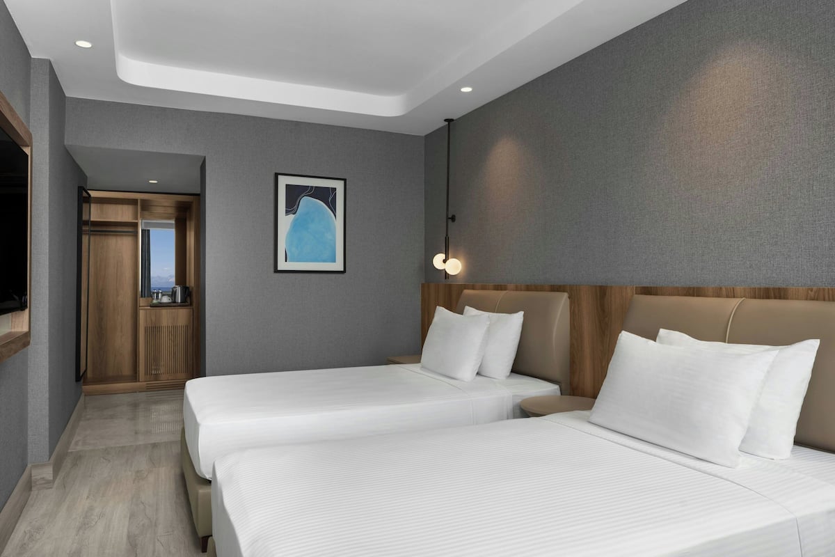 A hotel room with two beds, a wooden headboard, a framed artwork on the wall, and a view through an open door.