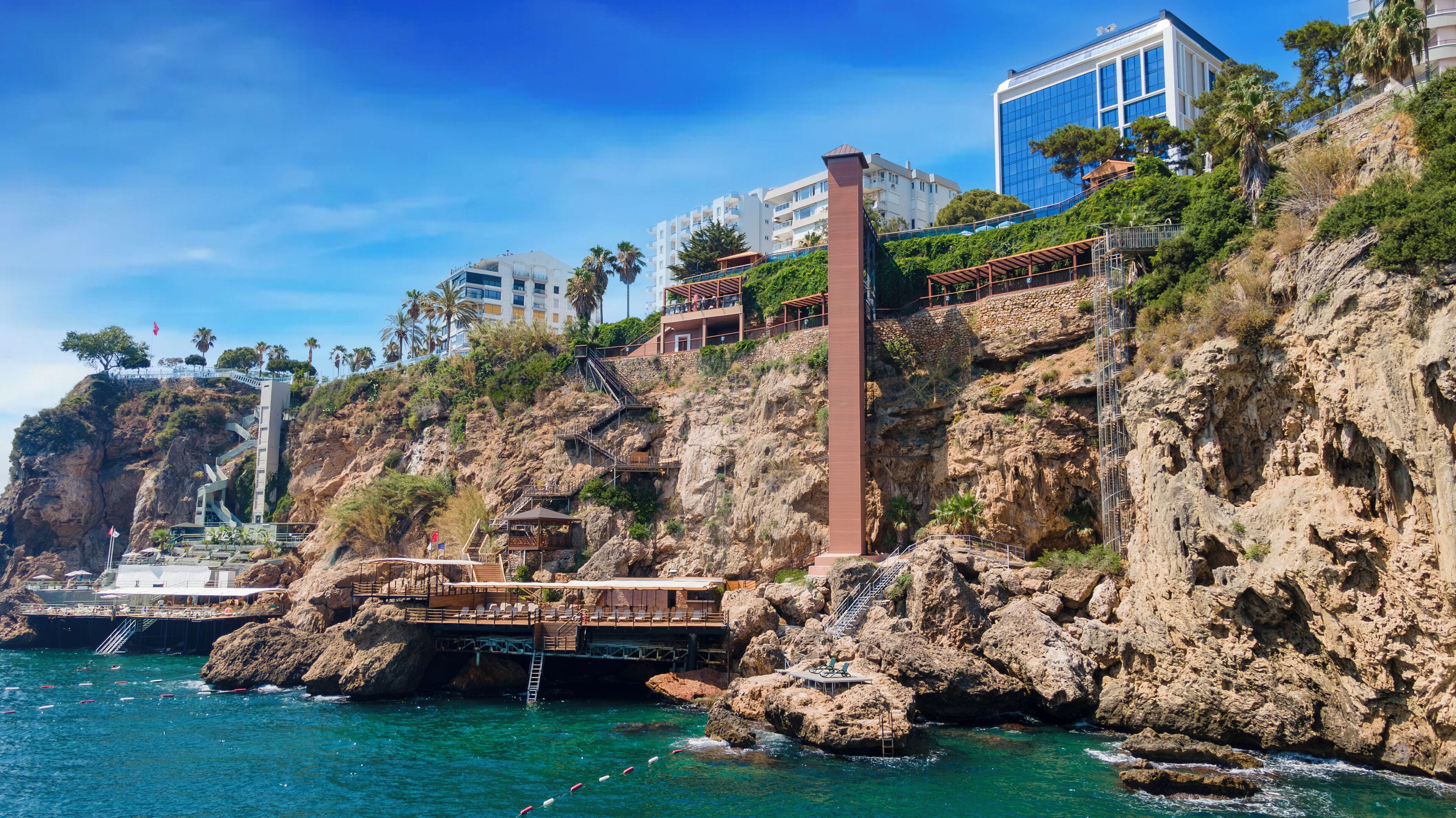 Un hotel costero con un acantilado rocoso, un puente que conecta con la playa, y un edificio moderno con ventanas de vidrio.