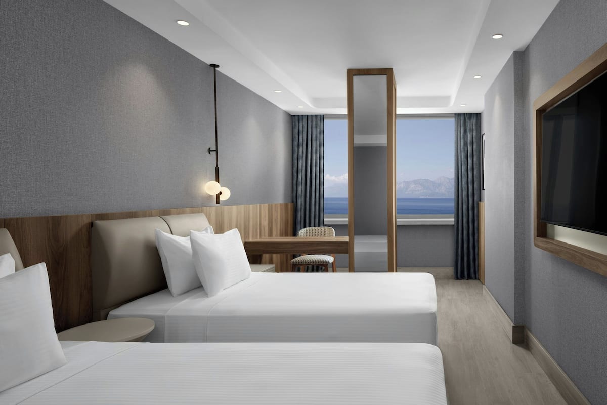 A modern hotel room with two beds, a desk, a chair, and a large window offering a view of the mountains.