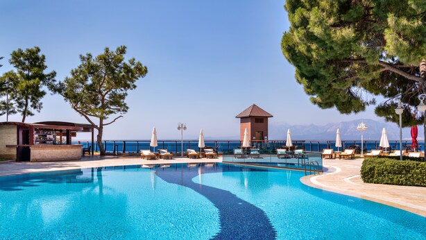 A clear blue pool with lounge chairs and umbrellas, a small building, and a view of the sea. - Delta Hotels Antalya Lara (Antalya)