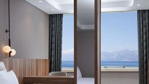 A hotel room with a bed, a desk, and a chair. There is a large window with a view of the sea and mountains. - Delta Hotels Antalya Lara (Antalya)