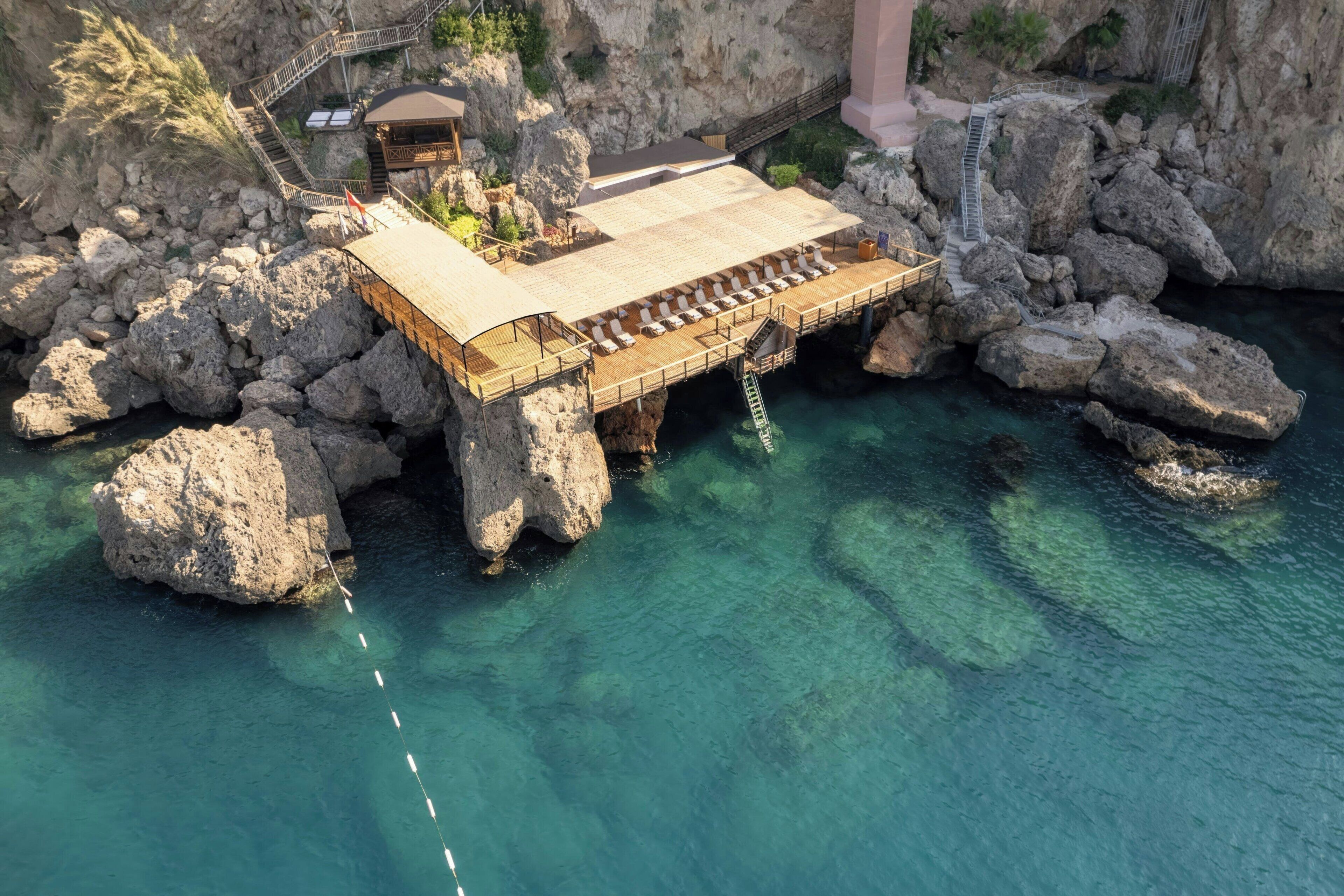 A wooden deck with lounge chairs extending over a rocky cliff into clear blue water.