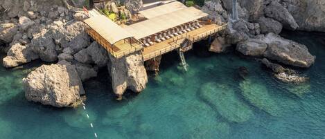A wooden deck with lounge chairs extending over a rocky cliff into clear blue water.