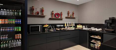 A modern kitchen with a refrigerator stocked with beverages, a microwave, and various food items on the counter.