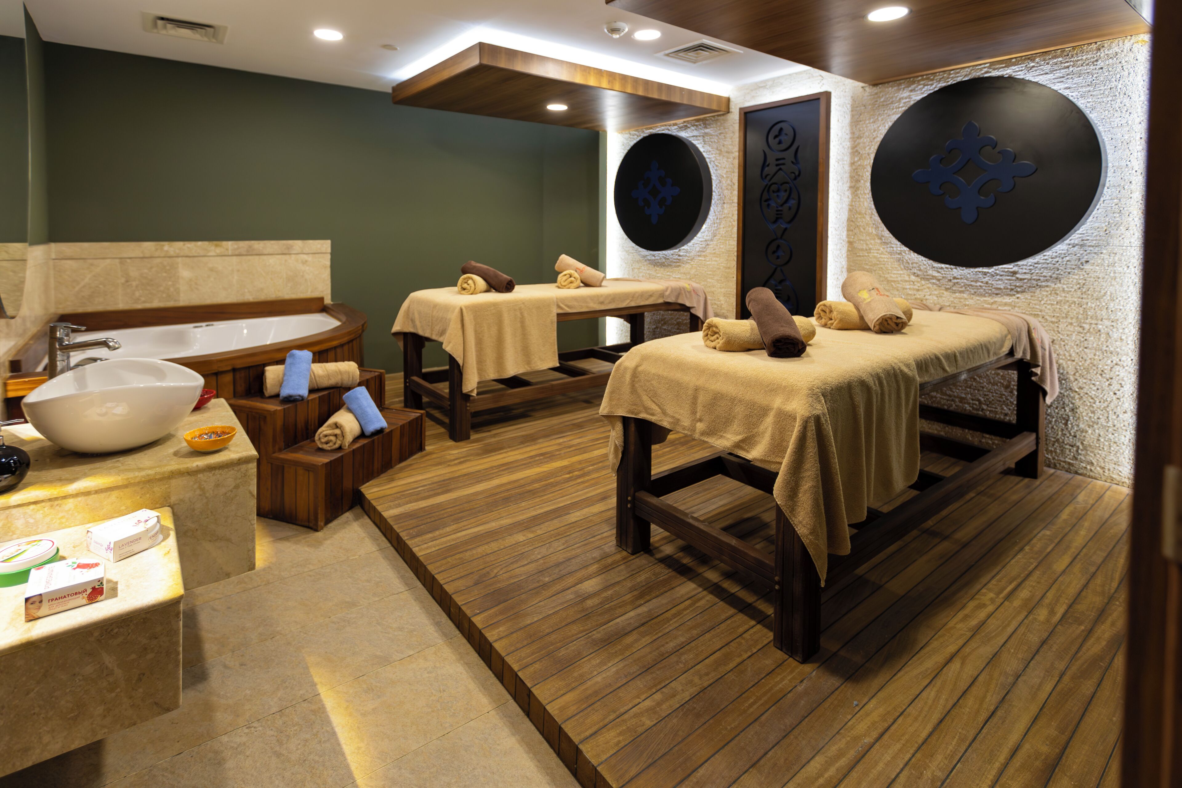Een spa-ruimte met een groot bad, twee massagetafels, houten vloeren en een wastafel.