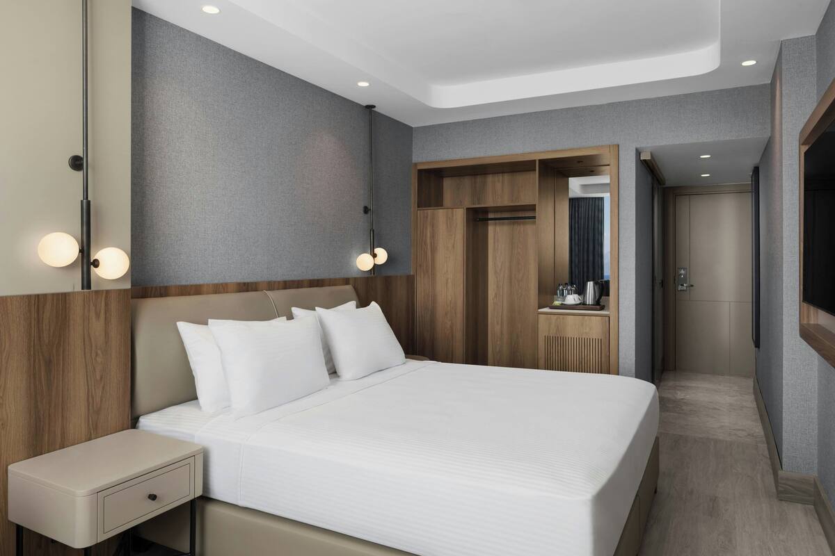 A modern hotel room with a large bed, a wooden wardrobe, a vanity with a mirror, and a small desk with a lamp.