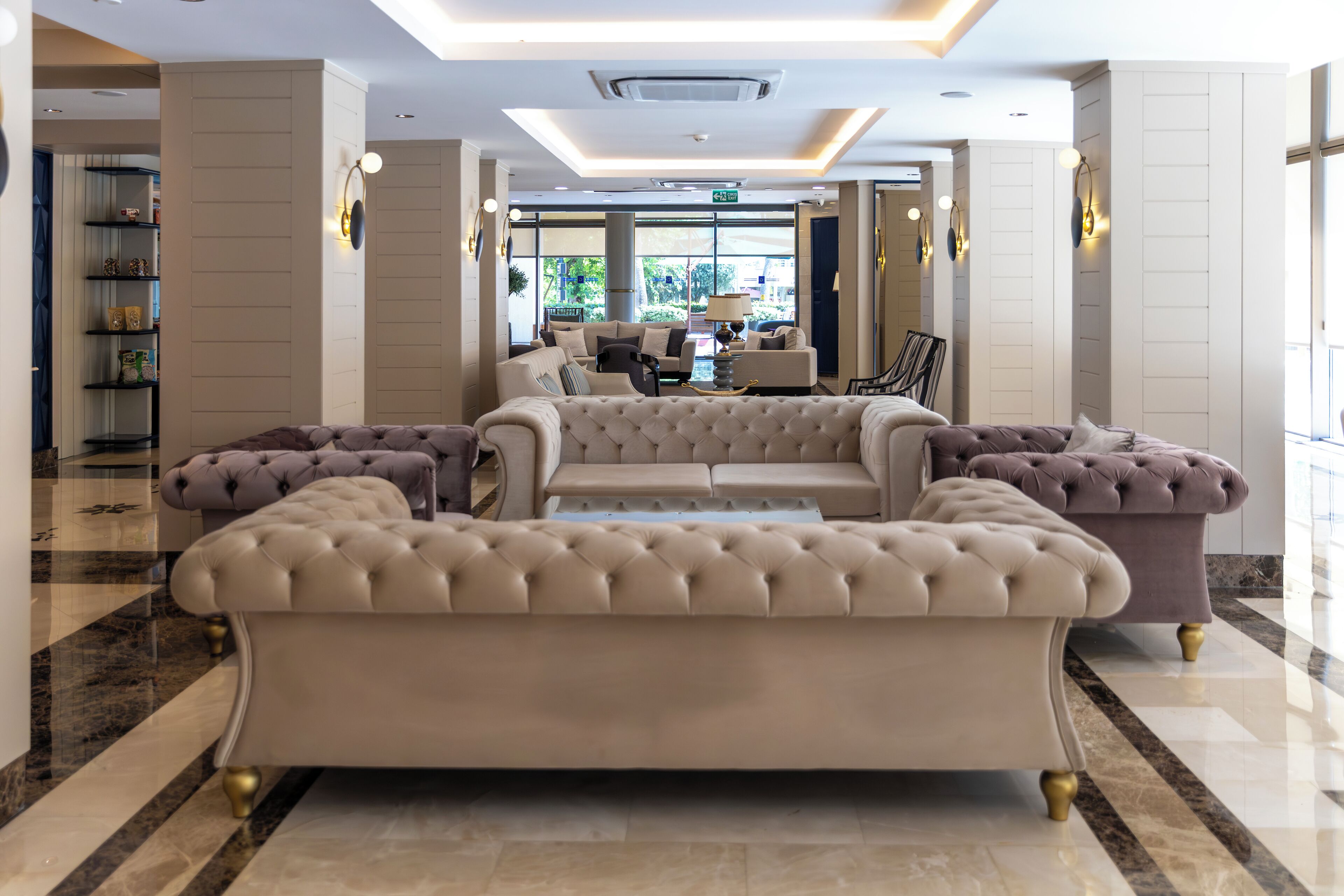 A spacious lobby with a large beige sofa, a smaller armchair, and a coffee table. There are shelves with decorative items and a large window providing natural light.