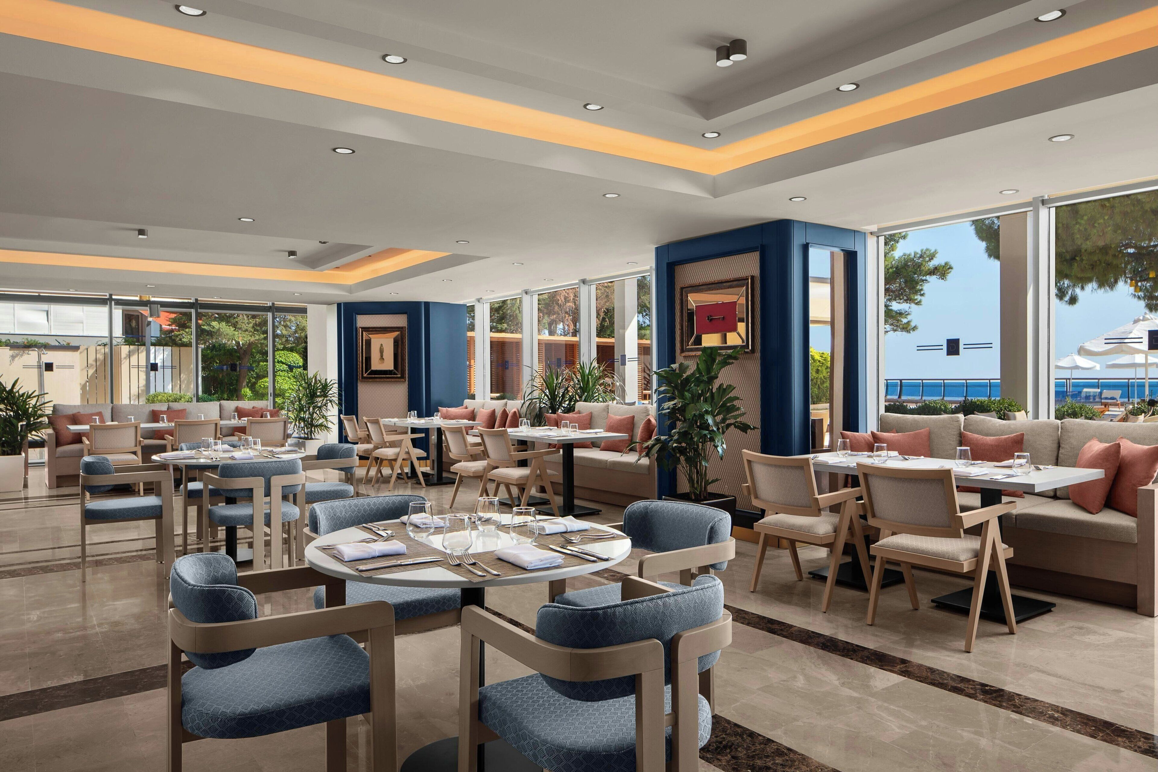 Een modern restaurant met ronde tafels en blauwe gestoffeerde stoelen, met uitzicht op zee door grote ramen.
