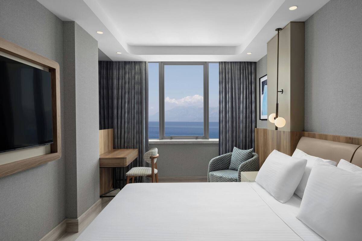 A hotel room with a large bed, a desk, a chair, a TV, and a view of the sea through the window.
